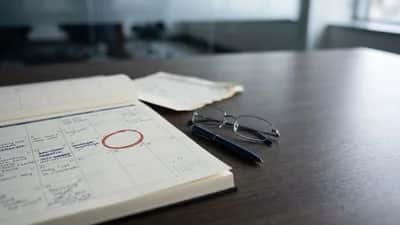 Zapisany planer na stole konferencyjnym, obok oderwana kartka, okulary i długopis; jedna data zaznaczona ceglastym kółkiem.